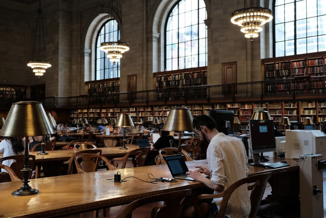 Man typing on a laptop, sitting at a wooden desk in a large study hall. The room is dimly lit, with wooden shelves full of books along the wall beneath large curved windows.