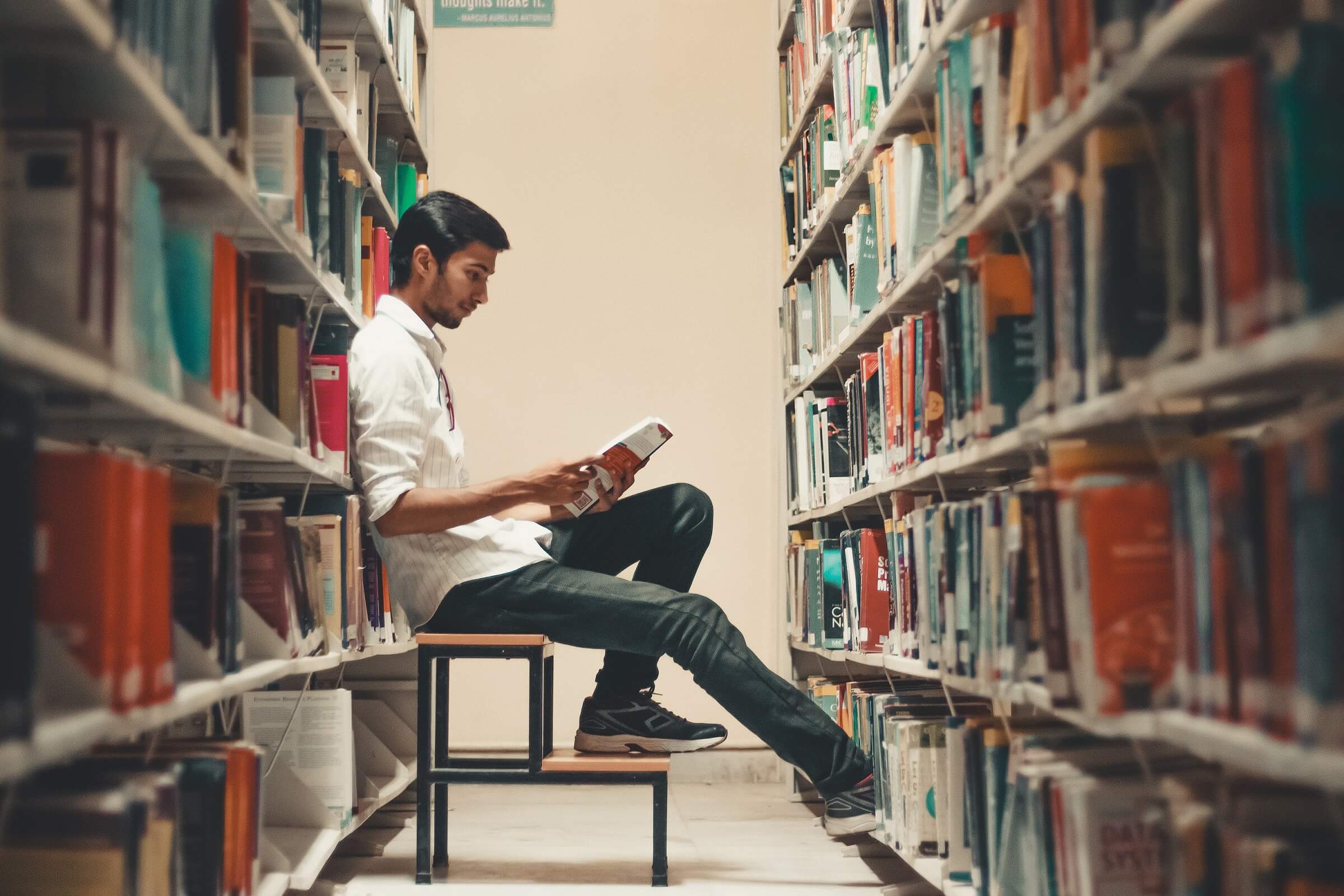 Man sitting on a step that is positioned in between two long bookshelves. He is reading a book.