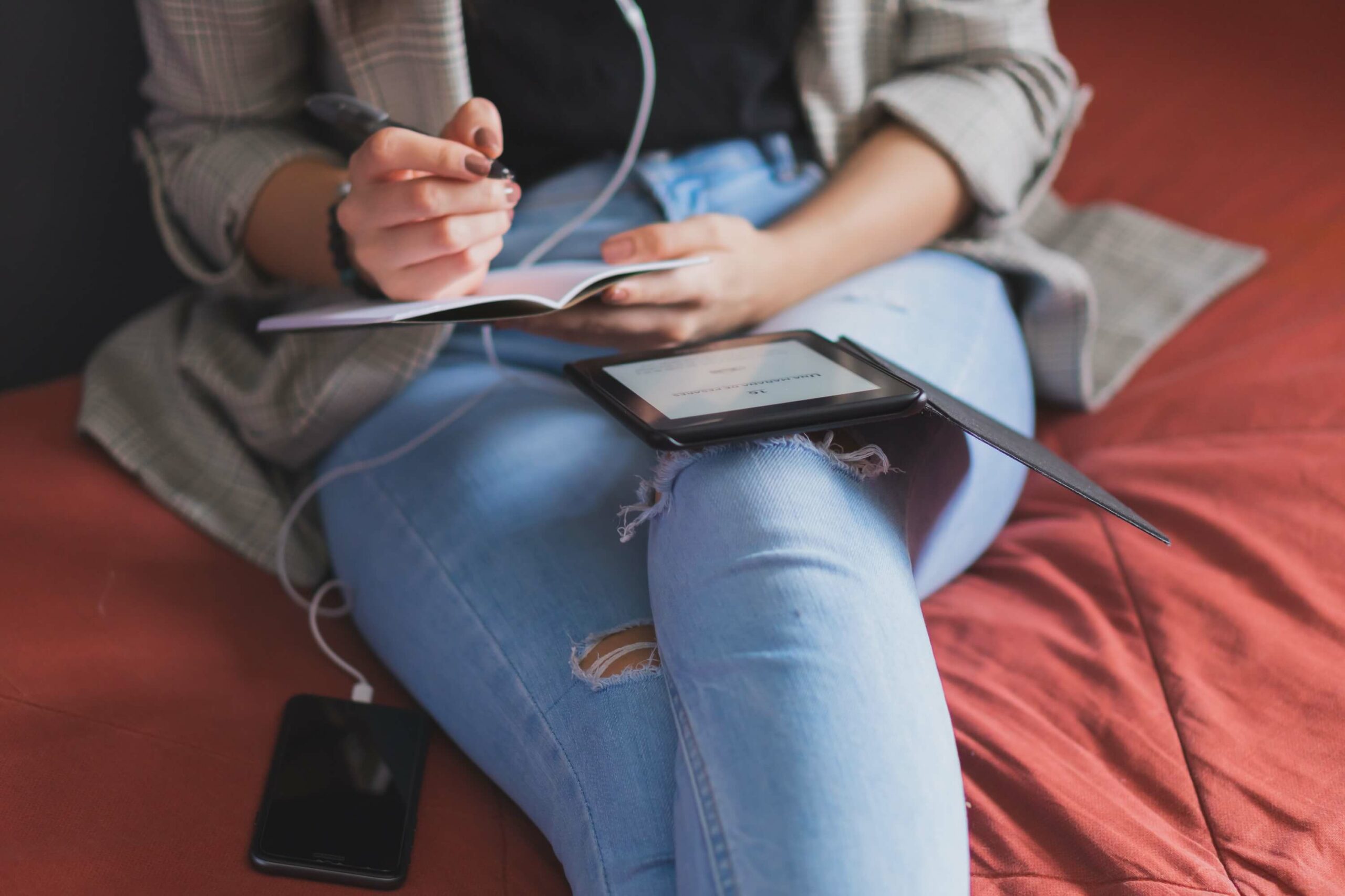 Woman writing in a notebook whilst sitting on a sofa. She has a kindle on her lap, and headphones plugged into her phone.