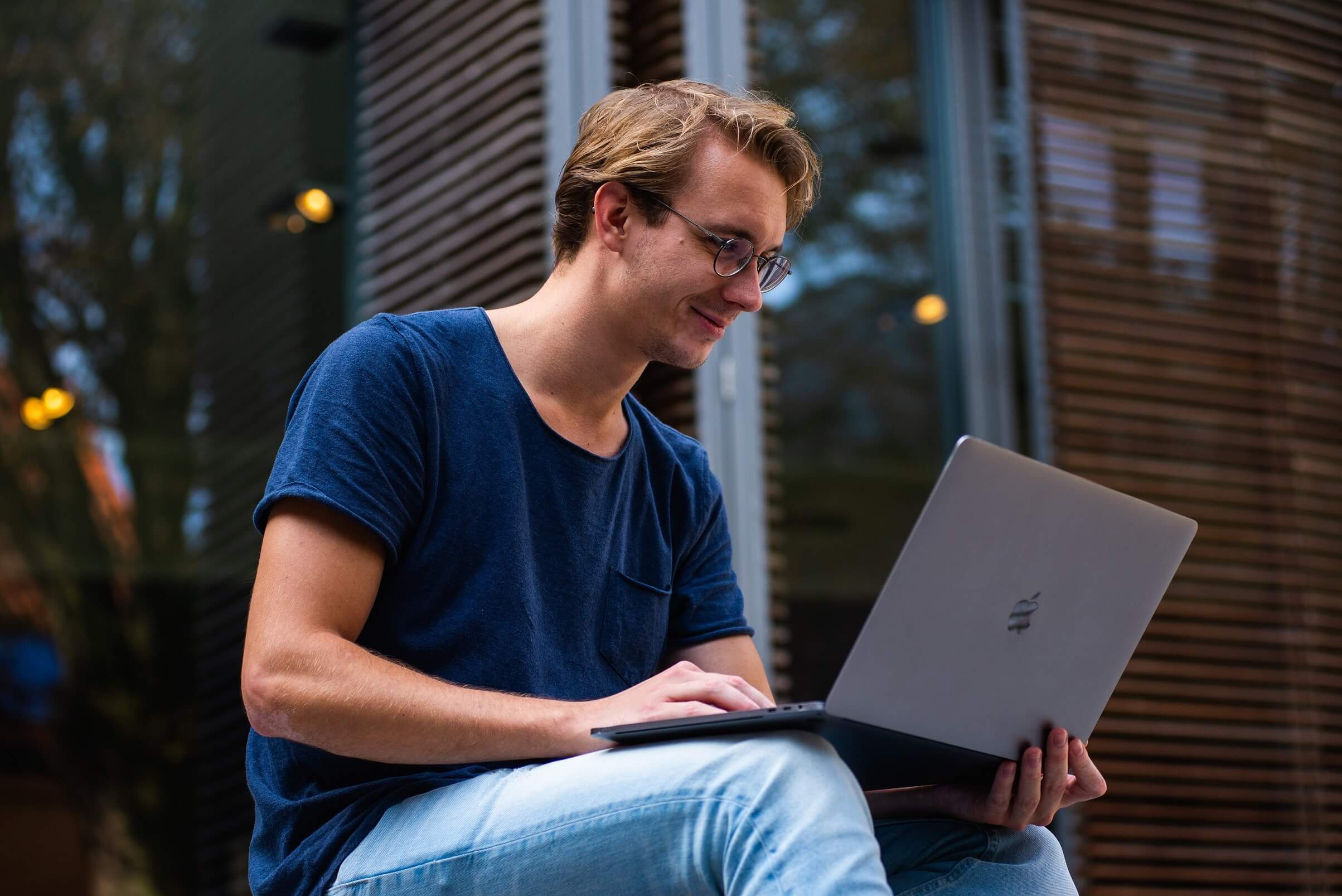 Person in blue t-shirt and blue jeans sitting down and working on laptop, smiling.