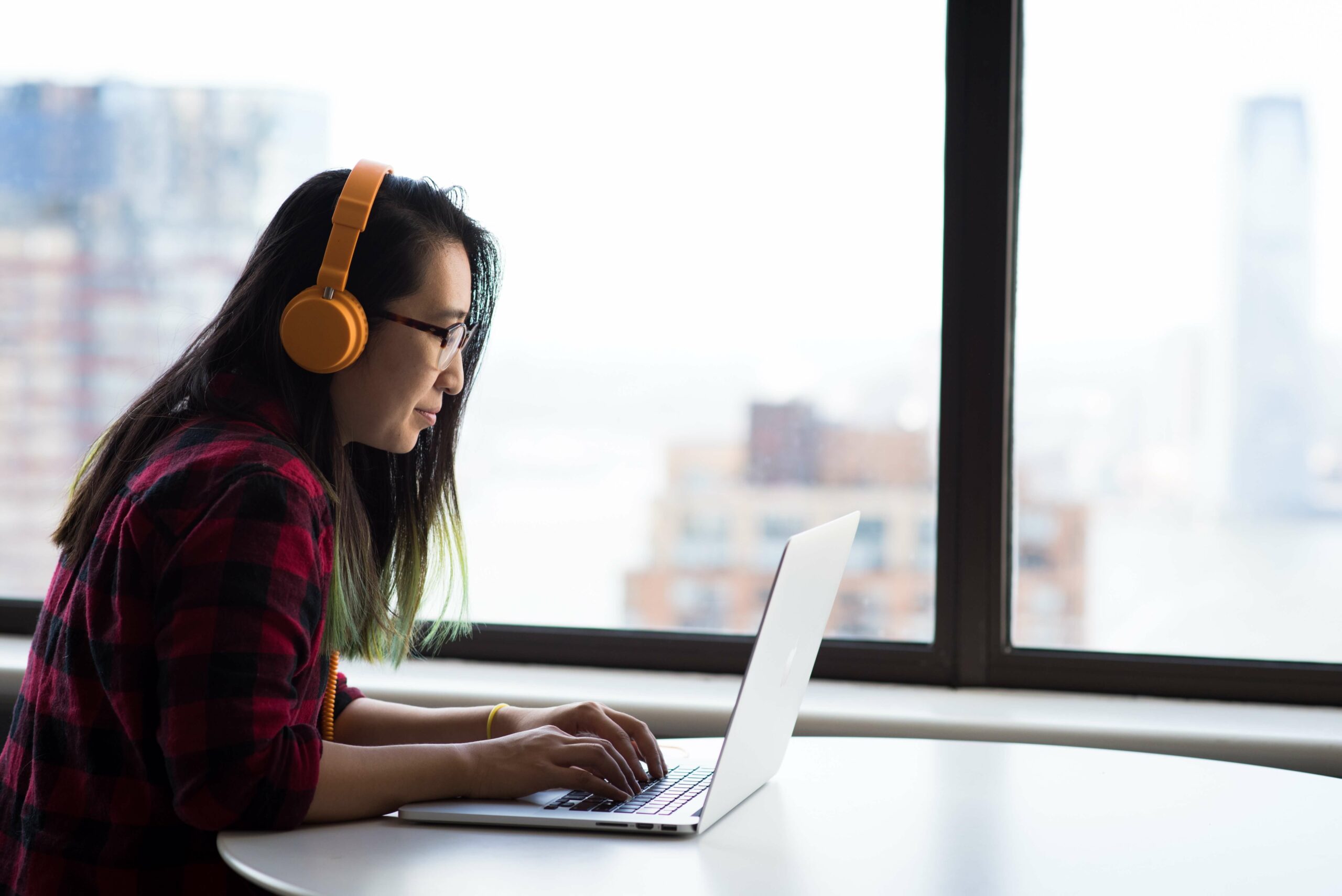 Person wearing orange headphones and a red and black plaid shirt, typing on a laptop at a desk in front of a large window.